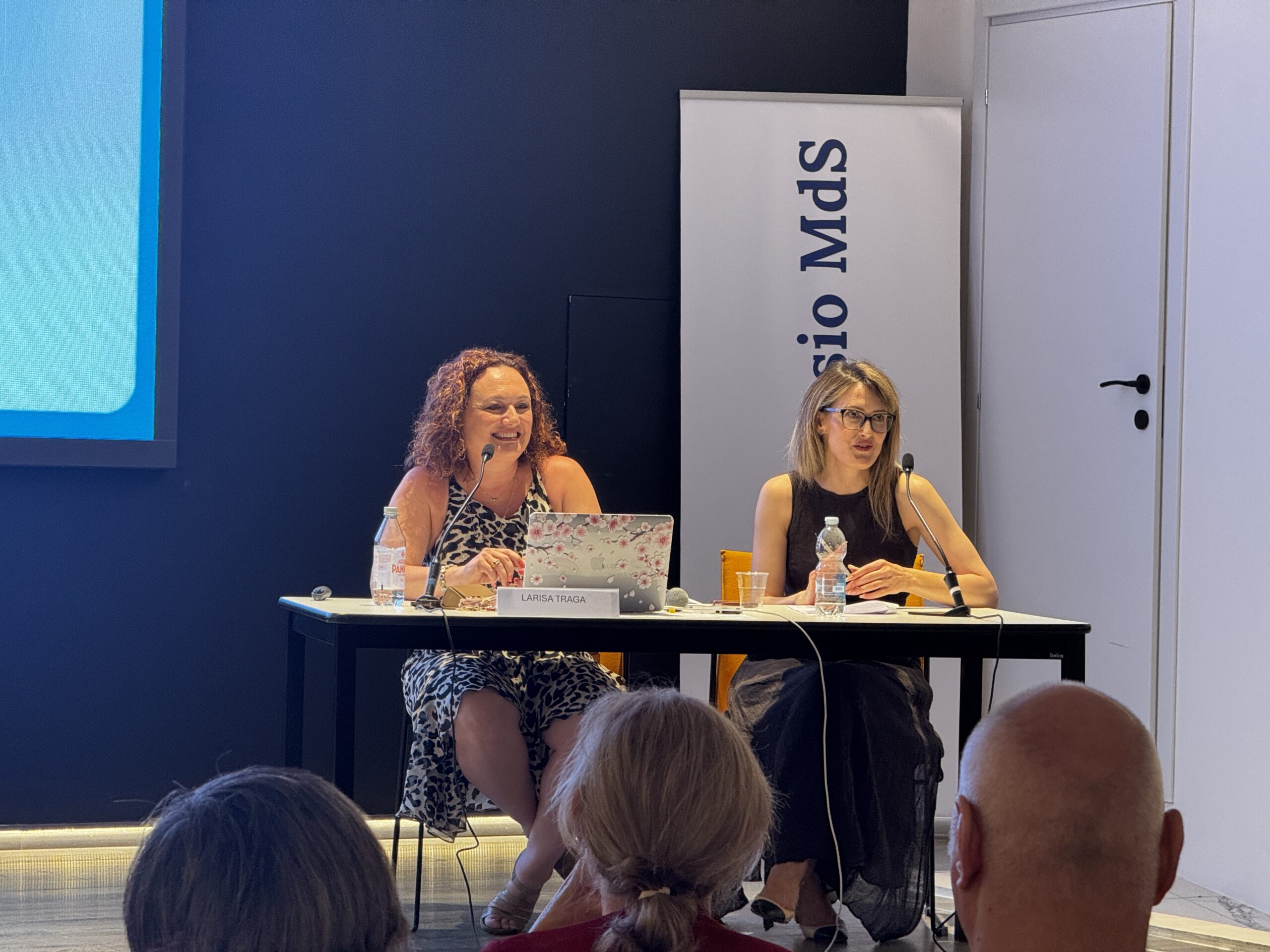 Larisa Tragaj e Laura Tragaj al tavolo dei relatori durante un workshop: Piano americano delle due relatrici sedute dietro a un tavolo bianco. A sinistra, Larisa Tragaj parla al microfono gesticolando con la mano destra; a destra, Laura Tragaj la ascolta con attenzione. Alle loro spalle è presente un roll-up aziendale con la scritta "S. Alessio MdS".