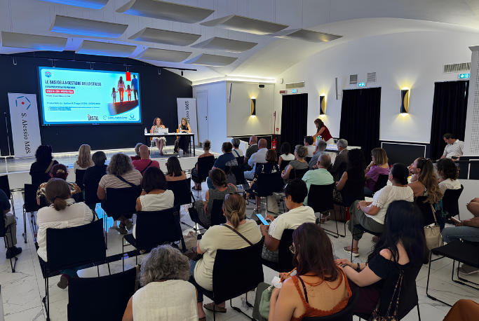 Workshop di formazione in sala conferenze: Vista da dietro della platea gremita di partecipanti seduti mentre assistono a una presentazione. Sul palco in fondo, due relatrici siedono a un tavolo davanti a un grande schermo che proietta le slide del corso intitolato "Le basi della gestione dello stress".